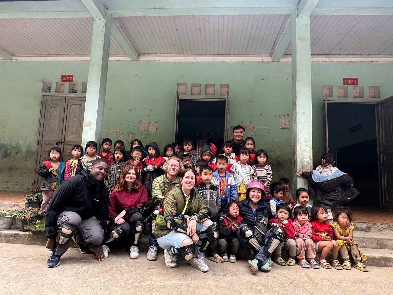 Foreigners sitting with local children smiling happily in front of an old small school. 