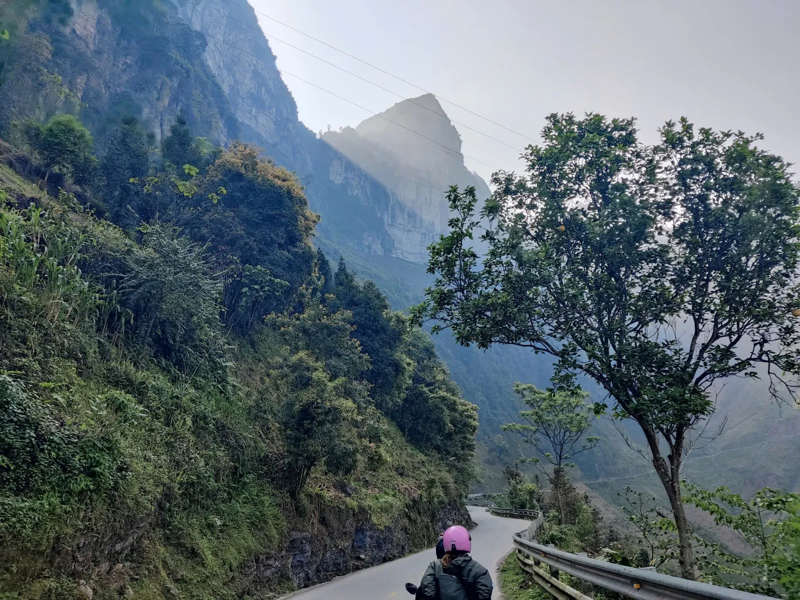 Motorbike travelers riding down a winding asphalt mountain pass surrounded by lush green valleys and dramatic karst slopes on the Ha Giang Loop